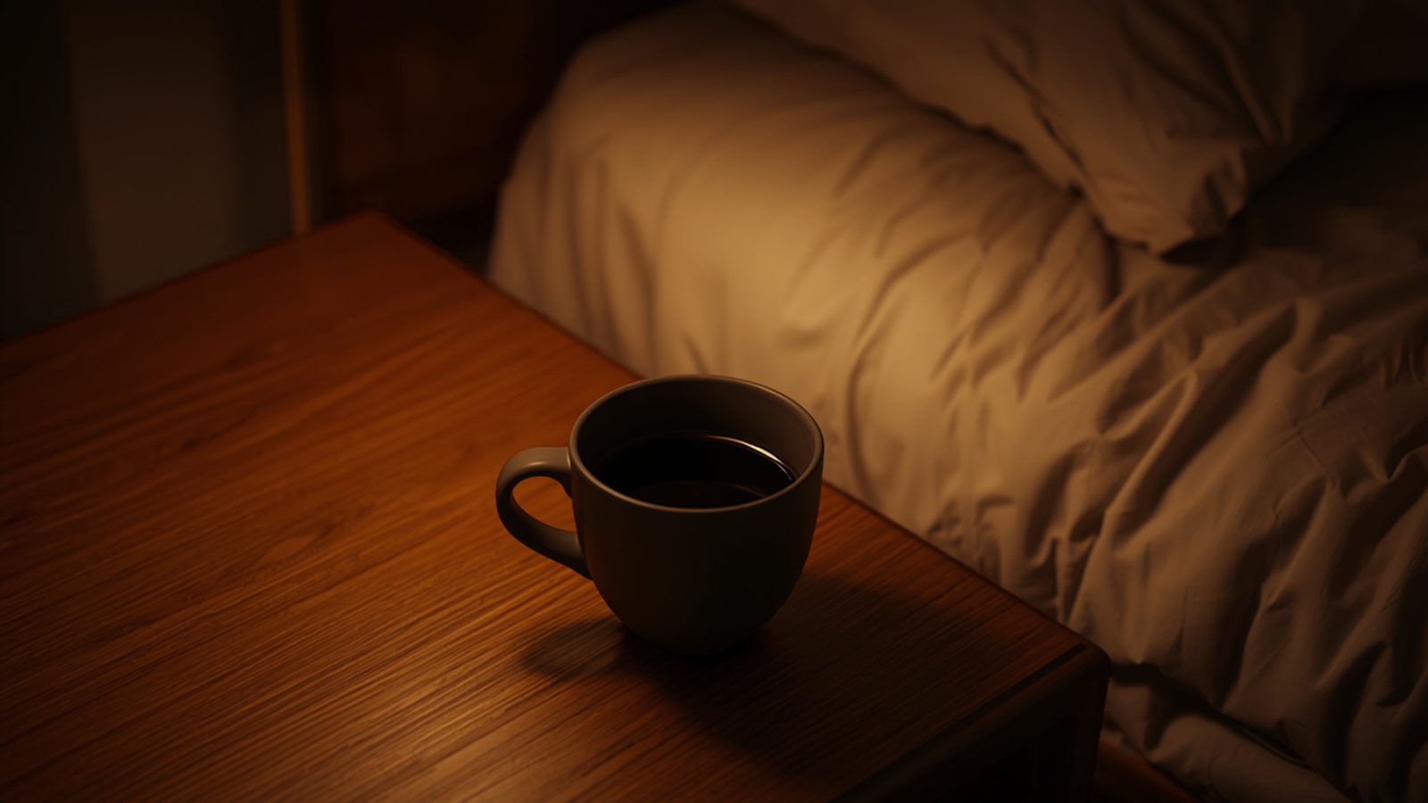 A cup of black coffee on a wooden bedside table beside an unmade bed in a dim, lamp-lit bedroom at night